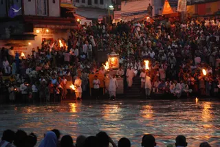 Evening Ganga Aarti at a riverside ghat with a very large crowd of devotees packed on the stone steps and the riverbank, many holding small flames. In the centre, several men in white dhotis and kurtas stand near a lit lantern-style shrine and wooden platforms, holding flaming torches and oil lamps over the water. A dark river reflects the golden lights. Buildings with signboards and a red-lit shrine line the background, and the lower foreground shows the backs of spectators.