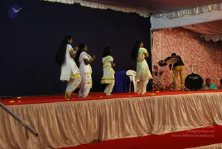 Four young women in white and cream traditional dress with gold-trimmed borders perform a dance on a raised stage with a red carpet and scattered yellow petals. Their long dark hair flows as they move. Behind them a navy backdrop and pink draped curtains frame a drum kit labelled BASIX, with a drummer in dark clothing at the right edge and a seated person near the drums. A white plastic chair sits on the stage. The setting is a festival stage under a white tent canopy.