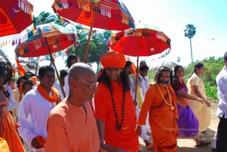 Outdoor procession on a dirt road under bright sky with palm trees, with red and multicolored decorated umbrellas with tasselled fringes held above the group. In the centre, a younger man in a saffron-orange robe and orange turban with a long dark rudraksha mala walks beside a bald older man in orange robes and glasses. Behind them, a bearded man in an orange robe wears a gold-beaded necklace, and women in purple and yellow saris and men in white shirts with orange shawls walk along. The younger central figure has long dark hair and shows the cues of a person candidate, though identity is uncertain from appearance alone.