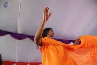 A person in a flowing saffron-orange garment stands under a white tent ceiling trimmed with purple and red drapes, one arm raised high with an open palm and the other hand holding the orange cloth outward. Long dark wavy hair falls past the shoulders, and the figure appears to be mid-gesture during an address or blessing. Only one physical person is visible.