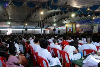 A large crowd seated in red plastic chairs under a white and blue draped tent at a convention. Many attendees wear white clothing and headphones; some wear yellow lanyards. A stage at the back shows a banner reading Nithyananda and a monitor displaying a figure in orange. Overhead lights and speaker stacks hang from the tent frame.