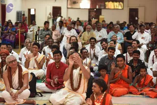 A photograph of a seated audience inside a hall during a satsang-style gathering. In the foreground, a bald man in white dhoti and shawl with rudraksha malas sits cross-legged, hands raised to his face. Beside him, a man in a red shirt with a rudraksha mala smiles, and on the right a young boy and a younger girl in saffron-orange clothes with beaded necklaces sit near the front. Behind them rows of seated men and women in white and light clothing clap and smile, with a woman in a purple sari visible at left. Gold-trimmed curtains and a framed picture on the wall are in the background.