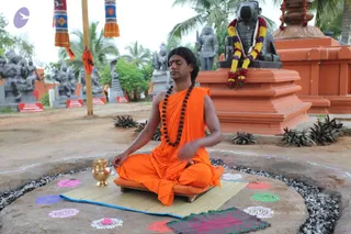 A seated man in saffron-orange robes meditates outdoors in a temple grounds setting, with his eyes closed, a long dark hairstyle, a rudraksha mala around his neck and a red tilak on his forehead. He sits on a low wooden platform on a straw mat, with a brass kalasha beside him. Behind him stands a large black stone Nandi bull draped with garlands of yellow, purple and red flowers, next to orange stone pedestals and grey stone deity murti figures among palm trees. Coloured rangoli designs decorate the ground around him, and red signboards line the background. Swamiji (probable) is shown in an appearance-based label, with his saffron robes, long swept-back hair and rudraksha mala as the cues.