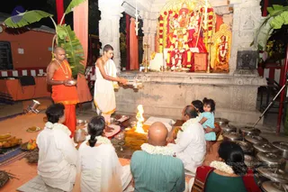 Evening ritual at a temple shrine. A fire burns on a raised stone hearth in front of an ornate shrine decorated with marigold garlands and holding a deity murti. A man in orange robes and a rudraksha mala stands at left holding a dish, and a young priest in white dhoti with a gold-trimmed cloth lifts a flaming aarti lamp toward the shrine. Seated devotees wear white with flower garlands, one woman in a red and green sari, and a small child in blue sits on a lap at right. Brass lamps and steel vessels are arranged around; banana leaves frame the scene.