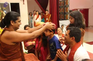 A man in an orange-brown robe with a rudraksha mala around his neck places both hands on the head of a boy in a blue t-shirt, pressing a pink-and-gold kalasha-like vessel down onto the boy's head as a blessing. Beside them a girl in a pink top with a mala stands with a similar pot. A smiling man in white with a red-orange shawl kneels with palms joined on the right. Behind them women in white and orange-trimmed clothes stand near red curtains, and a girl with long dark hair in a plaid top watches at the right. The setting resembles an ashram hall with a stage lighting rig and floral decorations at the left.
