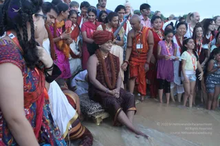 Outdoor beach event at the shoreline where shallow water washes over sand. A central seated figure wears a rust-red turban, a dark maroon wrap draped over the shoulders, a dense mala of rudraksha beads and a gold necklace, sitting on a leopard-print cushioned gold throne with bare feet in the surf. Around them, a crowd in orange, red, saffron and white clothing wears lanyards and watches, some holding a ceremonial red-tasselled staff and a long metal rod. A man on the right in an orange wrap and rudraksha mala gestures toward the seated figure. The figure is smiling, and the scene suggests a ritual beach visit with devotees gathered close. Approximately 20 to 30 people visible.