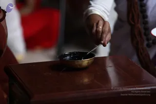 Close photo of a hand stirring a dark paste in a small brass bowl placed on a polished wooden table. The hand wears a thin gold ring, and a rudraksha mala with brown and dark beads hangs at the right edge beside a white-sleeved arm. A blurred red fabric and seated figures appear in the background. The paste may be an offering mixture used in ritual preparation, but the image alone does not show a specific ceremony.