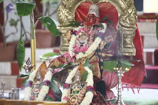 A photograph of a deity murti in a temple setting, seated before a gilded golden arch with a red cloth backdrop. The central figure is a red-faced goddess-like murti with a tall silver crown, a silver nose ring, and a pink and white flower garland with green leaves. Two smaller silver murtis stand in front, each draped with white jasmine garlands and pink or purple flowers. Silver oil lamps with lit flames, a silver tumbler and brass vessels sit on the altar, with banana leaves and pink petals scattered around. Potted plants and a red-and-white striped wall appear in the background. No people are visible.