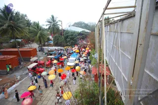 A photo of a procession on a paved path lined with palm trees, with a crowd of many people walking behind a white van decorated with a blue cloth. Numerous people hold colourful umbrellas in yellow, orange, red, pink and green, while others in white and cream clothes walk in the crowd. Red-and-white tractors with trailers and orange shipping containers stand along the left side, and a tall metal scaffold with a grey wall runs down the right.