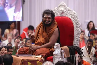 A smiling bearded man with long dark hair sits on an ornate silver-and-red throne, its back carved with a sun face and its arms shaped like mythical creatures. He wears a saffron-orange shawl wrapped over his shoulders and holds a wooden staff upright in one hand. A small table draped in orange cloth with a book sits in front of him. Seated audience members with a mix of white, red and saffron clothing fill the foreground and background, and a screen shows a partial image at top left. a person: long swept-back hair, saffron shawl and a throne-seated stance, presiding over a discourse or satsang.