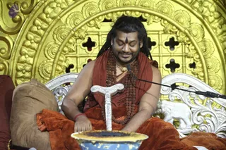 A seated man in saffron-orange robes sits on an ornate silver-and-gold throne with a gilded floral backdrop. He wears long dreadlocked hair, a vertical tilak on his forehead, multiple rudraksha malas and a gold chain, with a red thread on his wrist. He rests one hand on a silver ceremonial staff topped with a carved handle, and his other hand touches a blue-and-gold bowl in the foreground. A microphone stands beside him and a silver elephant motif decorates the throne. Appearance-based label: a person, seated on the throne with rudraksha malas and saffron robes, performing a ritual with the staff and bowl.