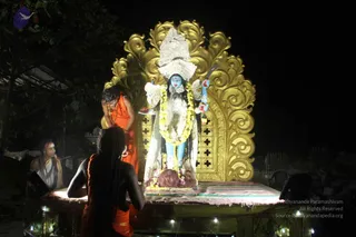 Night-time outdoor scene at a temple-like shrine with a large blue-skinned goddess murti, Kali iconography, with a white-and-blue face, protruding red tongue, white crown, yellow marigold garland and multiple hands holding objects. She stands before an ornate gold arched backdrop with a green base platform lit by string lights. In the foreground, a figure in orange robes with long matted hair stands before the murti, while another seated figure in white with a striped shawl and sacred thread on the left wears rudraksha beads and a forehead tilak. A third person with long hair in a dark garment kneels at the platform's edge. Trees and foliage frame the top left.