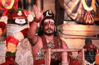 A photograph of a man seated on a wooden chair with a raised right hand in a speaking gesture, in front of a microphone on a stand. He has long dark hair with a crescent moon ornament and a red bindi, a white forehead band, a beard and moustache, and wears a leopard-print cloth with a dark red shawl and layered rudraksha malas with a gold pendant necklace. Behind him are a large shivalinga draped in red and white flower garlands with silver adornments, and a black deity murti. An orange-gold draped backdrop with a patterned cloth fills the upper background. A person on the right appears to be seated in red as well.