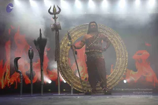 A figure stands on a stage in front of a large golden ring with carved relief patterns, backed by a red and orange fire-like projection and drifting haze. The figure has dark hair, a tilak-like mark on the forehead, bare arms with armlets and wristbands, a leopard-print tunic with a belt and a long rust-coloured dhoti. One hand grips a staff with a round finial and the other rests on the hip. To the left, a trident, a black ceremonial whisk, a curved hooked staff and a tall lamp-like pillar stand in a row. Stage lights shine from above.