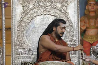 A photograph of a seated man with long dark hair, a white tilak on his forehead, a rust-orange robe over bare shoulders, and multiple rudraksha malas with a gold chain. He sits on a leopard-skin-draped seat before an ornate silver-repousse backdrop, pouring or offering from a small vessel into a shallow bowl in front of him. To the right, a large painted deity murti with orange and gold garments stands in the background. a person is seated in a ceremonial setting consistent with ritual worship.