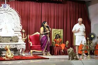 A stage event before a deep red curtain. A woman in a purple and gold sari with long dark hair and a jasmine-like flower in her hair speaks into a handheld microphone with her hands open. Beside her, a bald man in a white shirt and white shorts stands with a hand raised, wearing a headset. Behind them, a seated figure in saffron-orange robes, rudraksha beads and gold chains sits on a dark red draped platform with brass lion statues, and an ornate gold-framed backdrop is behind. A red velvet chair with gold trim, a white carved throne with a leopard-print cushion, a brass trident, a brass kalasha-style lamp stand and a white statue of a deity in a carved arch are also visible. Two brass oil lamps stand at the right edge.