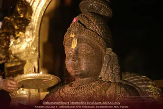 A dark stone or bronze-toned murti of a deity with a tall jatamakuta-style crown, a yellow vibhuti or kumkum mark and a small red bindi on the forehead, lit warmly against a black background. A gold-framed niche with ornate carving stands behind it. At the lower left, a hand holds a brass aarti plate with a small flame and a yellow-gold offering. The scene shows abhishekam to the murti in a temple setting, with the caption at the bottom reading Nithyanandeshwara Paramashiva Devalayam, Adi KAILASA, Navaratri 2021 Day 8 Abhishekam to SPH Nithyananda Paramashivam's Mula Vigraha.