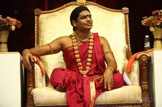A photograph of a man seated cross-legged on an ornate cream and gold upholstered throne with carved dark wooden armrests. He wears a red silk drape over his shoulder and torso, a red dhoti-style lower garment with a gold border, a heavy gold bead necklace, a silver-toned pendant chain, and gold armbands and bracelets. A red bindi marks his forehead and a microphone clip is visible near his face. An orange cloth lies over one armrest. Behind him are a dark red curtain and vases of pink and red flowers, with a water bottle on a side table.
