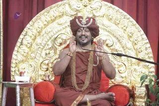 A seated man speaks into a microphone during a discourse, both palms raised in an expressive gesture. He sits on an ornate gilded throne with curved carved backrest, cushioned in red. He wears a maroon-red turban with a white jewelled ornament, a maroon-red robe, layered gold chains, a gold-bordered sash, bangles and a bracelet. Red drapes hang behind, and a small side table holds a glass and a plant sprig is at right. Probable Swamiji (appearance-based label): the candidate is seated in person on the throne with a crown-style turban, ornate ceremonial jewellery and ochre-red robes, and is teaching in a discourse setting.