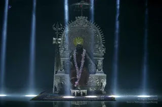 A seated figure on an ornate silver throne with a tall arched backrest, carved lion-like armrest figures and a silver trident leaning at its left side. The figure wears a golden crown, a long pink-and-white flower garland, and a reddish-maroon cloth, with bare arms, and sits on a purple patterned rug before a dark stage. Vertical blue light beams and floor spotlights illuminate the scene, and the reflection shows on the glossy floor.