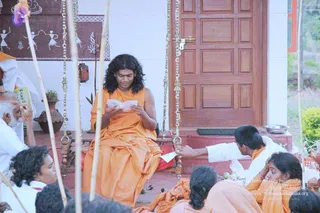 A photograph of a seated man in saffron-orange robes with long, swept-back dark hair, reading a small booklet on a raised red-tiled step in front of a wooden door with brass chains. Beside him, a bearded elder in white holds a small book and a younger person in orange sits on the step, while several people in orange and white sit on the ground in the foreground. A painted wall with folk figures and a clay pot with a plant are visible, with a few bamboo poles in the frame. a person: long swept-back hair and saffron robe, seated reading in the centre, consistent with the page's description of a monastic training session at an ashram.