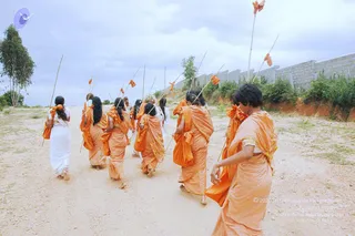 A group of women walks away from the camera along a dusty dirt path, carrying tall bamboo staffs topped with orange cloth streamers. Most wear orange saris draped over the shoulder, and a few wear white garments. Their hair is long and dark, and some tie orange ribbons at the hair. Behind them stands a grey concrete block wall beside green shrubs and a tall tree under a cloudy sky. The people are barefoot, and the scene looks like a monastic training activity at an ashram site.