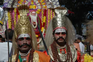 Two costumed performers stand in a procession in front of a flower-decorated float. Both wear ornate gold crowns shaped like a hooded cobra, with long pale hair falling beside their faces. Their faces are painted with white forehead bands, red eye and cheek markings and black upturned moustaches. The left performer wears an orange garment with a gold collar, white pearl necklace and rudraksha mala; the right wears red and gold armour-like chest plates with a rudraksha mala. Each holds a long blade or spear. Behind them a painted portrait of an elderly bearded man with white hair is set among garlands of yellow and orange marigolds and pink flowers. Onlookers stand in the background.