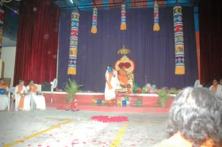 A stage event at an ashram hall, with a central raised golden throne (peetha) draped in red and decorated with flowers. A seated figure in orange clothing sits on the throne beside a microphone stand, beneath a small golden canopy. In front, a standing person in white with an orange shawl holds a blue balloon, and colourful balloons are scattered across the stage steps. Four or five women in white clothes with orange shawls sit at left and right of the stage, some smiling. Rose petals are scattered on the floor, tall decorative banners in red, blue and yellow hang behind the stage, and potted palms flank the stage. A person's head is visible in the lower right foreground facing the stage.