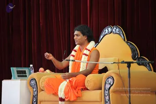 A photograph of a seated man in saffron-orange robes with a white and red flower garland draped over his shoulders, speaking with one hand raised on a gilded ornate yellow throne with carved armrests. Behind him hangs deep red velvet drapery, and a microphone on a stand is positioned in front. A digital clock display, a water bottle and a small plate of apples sit on a white table to the left. a person: the seated man has long swept-back hair and saffron robes with a flower garland, matching the page's Sarva Darshan event at Adi Kailasa, Bengaluru, though the face is not used for identification.