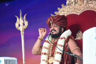 A seated man on a red-and-gold throne speaks into a microphone during a satsang. He wears a maroon turban with a gold crescent ornament and a small gold emblem, a maroon shawl over his shoulders, and layered white and red-flower garlands with gold beaded necklaces. He has gold bangles and an armlet, and his raised right hand is curled in a gesture of teaching. Beside him stands a tall gold trident. The blue-to-magenta backdrop and a white stage monitor complete the scene. Swamiji (probable): long hair is not visible, but the turban, crescent ornament, saffron-maroon ceremonial clothing, garlands and gold jewellery are consistent with the page.