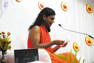 A photograph of a seated man in a saffron-orange robe with long, swept-back dark hair, reading a small pink card held in both hands at a stage. Behind him is a white backdrop decorated with red and yellow swirl motifs. A microphone stand curves in from the right, a flower arrangement of orange blooms sits at left, and a black monitor or laptop rests in the foreground. Probable Swamiji (long swept-back hair, saffron-orange robe) appears seated at a stage event. One person is visible.