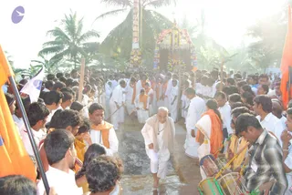 Outdoor procession on a wet path lined with palm trees under bright sunlight, with a decorated chariot or palanquin with red and gold canopy in the background. A central older man with white hair and glasses, wearing a white dhoti and white shirt with a light shawl, walks barefoot through the water-soaked path, flanked by a crowd of devotees in white clothes and saffron/orange shawls. Drummers in the lower right play drums, and tall orange flags and white flags with a blue emblem line the sides. Many people are visible, numbering over fifty.