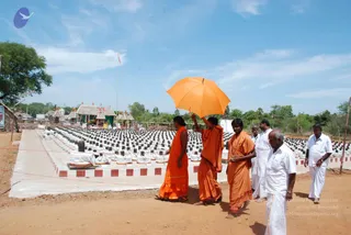 Outdoor photo at a temple grounds with a large open courtyard lined with rows of black Shiva lingams on white-draped bases. Four people in saffron/orange robes walk along a concrete path; the central figure holds up a bright orange umbrella over another saffron-clad person. Several men in white dhotis and shirts walk behind them on red-and-white kerbed edges. Thatched-roof shrines and a tall flagpole stand in the background under a blue sky with clouds and trees.