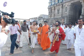 Outdoor photo on a paved stone path before a tall, ornately carved stone temple gopuram with painted tiers. A group walks toward the camera in a procession. In the centre, a bare-chested man in white dhoti carries a bundle of bananas. Beside him, a bearded man in saffron robes walks with a hand raised. At right, a bare-chested bearded man in red cloth carries a kalasha-like pot wrapped in plastic, with a garland of yellow flowers hanging from his arm. Men in white shirts and white dhotis surround them, some wearing rudraksha-style beaded necklaces. At left, a video camera operator films the procession.