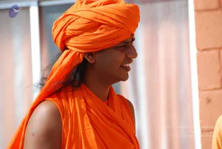 Close profile photograph of a smiling man wearing a bright saffron-orange turban and matching saffron-orange sleeveless wrap over his shoulders, with long dark hair falling from beneath the turban and a small red vertical mark on his forehead. Behind him are pale vertical panels and a brick wall edge at the right, with a partly visible person in yellow at the far right edge.