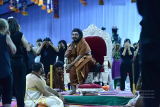A bearded man in an orange-brown shawl and rust-coloured dhoti sits cross-legged on an ornate silver throne with a red velvet seat, holding a handheld microphone in his right hand and speaking. Below him on a green-and-red striped mat, a footstool stands before the throne. A seated man in a white dhoti with a gold border and a bare-shouldered upper body sits to the left, and a man in rust-coloured robes with a rudraksha mala sits nearby. Behind them, women in black and purple clothing press their hands over their eyes in a meditative gesture, while tall yellow poles and hanging orange-and-yellow tassels decorate a blue-draped hall.