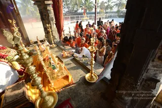 A photograph taken from an elevated vantage point inside a stone temple hall. A bald man in white ritual clothes bends over a stone platform, his hands reaching toward a brass plate of offerings in front of a gilded deity murti set on an orange cloth. Around him, a crowd of devotees in orange and red robes, some with garlands, stand with palms pressed together in namaste. Tall brass oil lamps stand in the foreground beside trays of red flowers, fruit and sweets. Carved dark stone pillars frame the scene, and an outdoor courtyard with palm trees and a golden shrine is visible through the opening. The figure in white could be a priest, but his face and appearance do not clearly establish that he is the a person, so the identity remains unconfirmed.