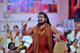 A photograph of a bearded man seated on a red throne-like chair in a hall, raising a golden arrow-shaped stick in his right hand while his left arm rests on the armrest. He wears saffron-orange robes with a matching draped shawl, a gold chain necklace, a rudraksha-style bead necklace and a red-and-gold wristband; long dark hair falls past his shoulders and a red bindi marks his forehead. Behind him stands a silver trident on a pole and a golden bird-headed ceremonial staff sits at his lower left. Seated devotees in white and saffron clothing fill the background, along with a hand-held fan and colourful abstract paintings on the walls.