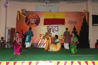 A stage performance at a hall with an orange and yellow backdrop reading 2010, decorated with marigold garlands, balloons and a silhouette of a temple tower. A circular portrait on the backdrop shows a smiling man in an orange turban and orange robe, labelled as a portrait, not a physical person. A yellow and red flag hangs centrally. Six performers stand and kneel on green stage matting: a girl in magenta and a girl in green saris holding small lamps, a boy in teal kurta and white dhoti, a boy in grey kurta, a woman in yellow sari kneeling with hands together, and a figure in gold and orange costume with a crown bowing. Red and white bunting edges the stage.