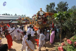 Outdoor procession on a dirt path beside a tin-roofed building and flowering shrubs under bright sky. A decorated palanquin with gold canopy, red and white garlands, tassels and a central kalasha carries a seated figure in red and gold. Men in white dhotis and shirts walk alongside, some holding red flags; a young monk in saffron/orange robes stands at left. Women in saris, including one in pink and one in blue, watch from the right. Colourful umbrellas and a small framed emblem are visible. The palanquin figure's face is not clearly visible, so no identity is asserted.
