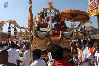 A golden palanquin procession moves through an outdoor ashram grounds under a bright blue sky. A seated figure in a red garland and saffron cloth rides beneath an ornate gold canopy with tassels and a trident finial, surrounded by garlands of white flowers. Men in white dhotis and orange shawls walk alongside and carry the palanquin, some wearing rudraksha malas and gold-trimmed cloth. Devotees line the foreground, and bright yellow and red ceremonial banners flank the scene.