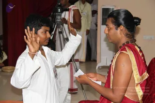 A young boy in a white shirt and white dhoti raises his right hand with palm open while facing a seated person in a red and gold silk garment with a rudraksha mala draped over the shoulders and a hair bun. The seated person holds a folded paper in both hands, appearing to administer a vow or oath. A cameraman with a tripod films from behind in the background, with a man in white dhoti and a yellow shirt nearby, a maroon curtain behind, and a white pedestal fan on the right. The seated person's appearance does not clearly match distinctive a person cues, so identity is unsure.
