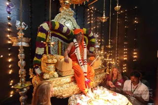 A photograph inside a decorated shrine room during a padapuja ritual. A man in saffron-orange robes and an orange turban sits on a gilded throne with a golden lion-shaped armrest, draped in a multicoloured flower garland and wearing a white and orange garland. He rests his bare feet on a tray of offerings. Brass oil lamps, hanging lamps and string lights surround the throne against a dark backdrop. Two visitors kneel at the lower right: a woman with light hair in a red patterned top and a man in a white shirt with a lanyard. A third person's hair is visible at lower left. Printed text in the corner reads Source , which is omitted from the text field