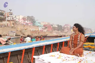 A photograph on a Ganga boat in Varanasi. A long-haired man in orange-saffron robes, a rudraksha-style white bead garland and gold armbands sits cross-legged on a white cloth strewn with marigold petals at the bow, smiling toward the riverbank. Several women in colourful saris (red, pink, teal) and a man in a white shirt sit in the boat behind him, and a boatman in pink rows on the left. Ghats, multistorey buildings and hazy sky line the river.