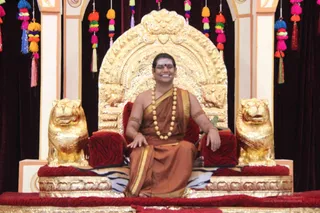 A smiling man in saffron-orange robes sits on an ornate gilded throne backed by a carved golden arch, flanked by two gilded lion sculptures. He wears a gold-bordered orange shawl, a long beaded necklace of wooden beads, gold armbands, and a red tilak on his forehead. The red-upholstered seat is draped with a striped tiger-print cloth. Colourful tassel garlands in pink, orange, green and blue hang behind the throne against dark red drapery.