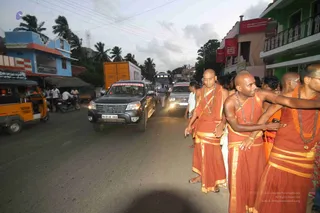 A street procession on a town road in Tamil Nadu at dusk. Several shaved-headed men in saffron-orange dhotis and shawls, with rudraksha and bead malas, walk along the roadside, some with hands extended. A dark Ford SUV with a NITHYANANDAM placard on its windscreen and a white van follow in the traffic. Green and pink shopfronts, palm trees, an auto-rickshaw and motorcycles line the road.