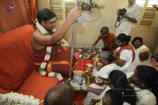 A photograph of a seated man in an orange-and-maroon ceremonial wrap with a long white floral garland and a red-and-white flower garland, holding a tall silver ritual staff with a round silver top. A tilak is on his forehead and he wears a rudraksha mala. Around a low table with brass plates, bowls and a golden kalasha, several women in white saris with red borders and a shaved-head man in orange sit on the floor, one holding a small vessel. A cameraman in white stands behind. Orange drapes hang behind the seated figure. a person appears to be the seated man in orange-and-maroon with garlands, long hair and rudraksha mala, presiding over the puja. The scene shows a home puja with women offering items on plates.