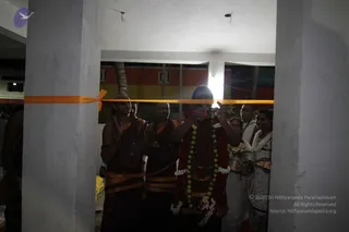 A photograph of a ribbon-cutting moment at a doorway inside a building with plain white pillars and a whitewashed ceiling. A man in a dark reddish robe and a heavy garland of yellow and white flowers stands at the centre, raising both hands near a bright orange ribbon stretched across the opening. Several monastic men in saffron-orange robes with rudraksha malas stand to his left, one smiling and clapping. Two women in white saris stand at right, one holding a brass plate with flowers and offerings. Behind the doorway is a multicoloured wall of orange, yellow and red with a palm tree visible.