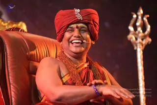 Photograph of a smiling man seated on an ornate red leather throne-style chair with gold trim, wearing a red turban with a silver brooch and a red bindi on his forehead, a red and gold garment draped over his shoulder and a long rudraksha-style mala necklace. He wears a small microphone headset and a blue beaded wristband. A tall golden trident stands at right against a dark red backdrop. a person is shown in person, appearing in a satsang-style setting. One person visible.