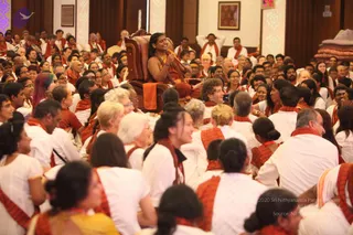 A photograph of a large seated gathering in a hall with wooden panelling and framed pictures on the walls. In the centre, a figure with long dark hair and an orange-saffron garment sits on an ornate carved wooden throne with an orange cloth draped over its arm, a rudraksha-style mala visible at the chest. Around them, many devotees in white clothes with orange-red shawls sit close together, some smiling and looking toward the throne. Stacked folded cloth sits at the right edge. Probable Swamiji is seated on the throne, identified by the long hair, saffron clothing and rudraksha mala. The crowd appears to be roughly 100 people.